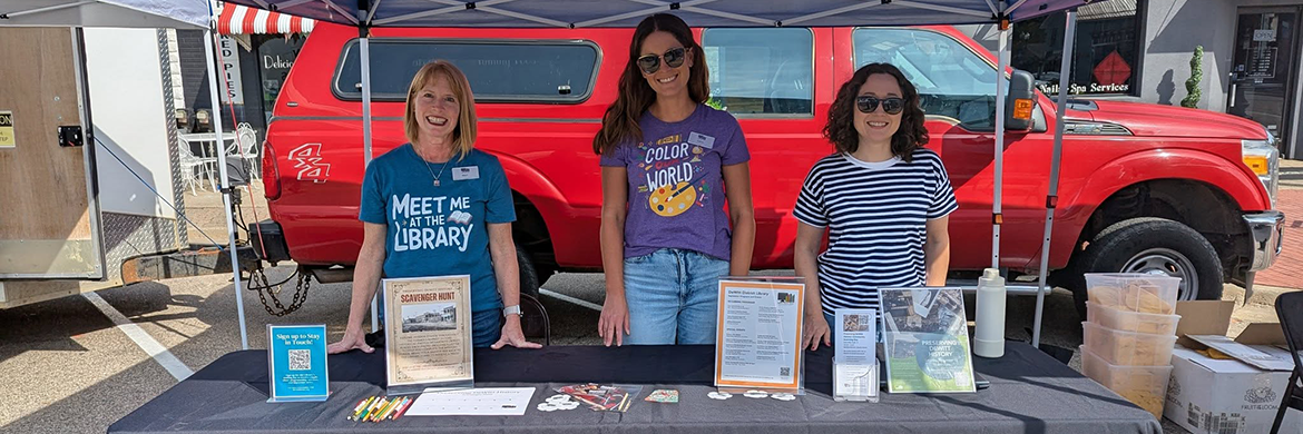 Three librarians smiling as they stand behind a table with the library logo during a community event
