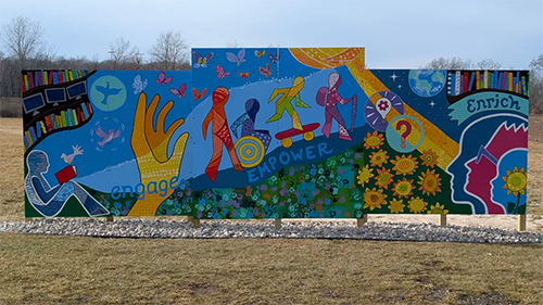 Large six piece canvas mural outside. The mural shows books, a hand reaching out for more, people adventuring outside, and silhouettes of people looking up and enriching their minds.