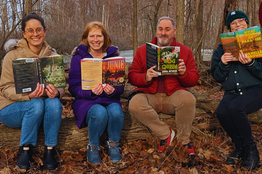 Group of four adults sitting in the wilderness with open books and smiling at the camera