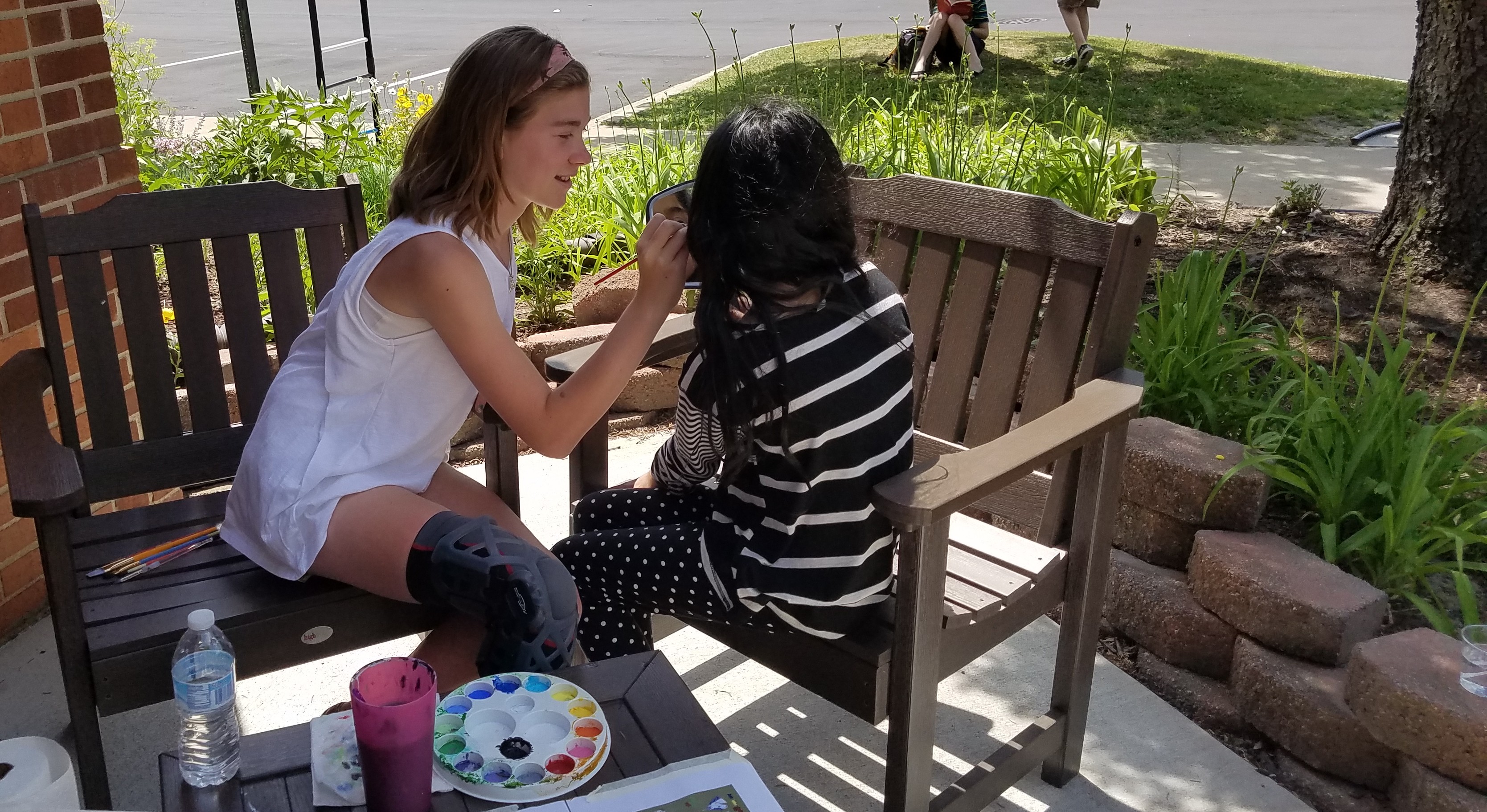 teenage girl sits outside with another young girl and paints her face with facepaint