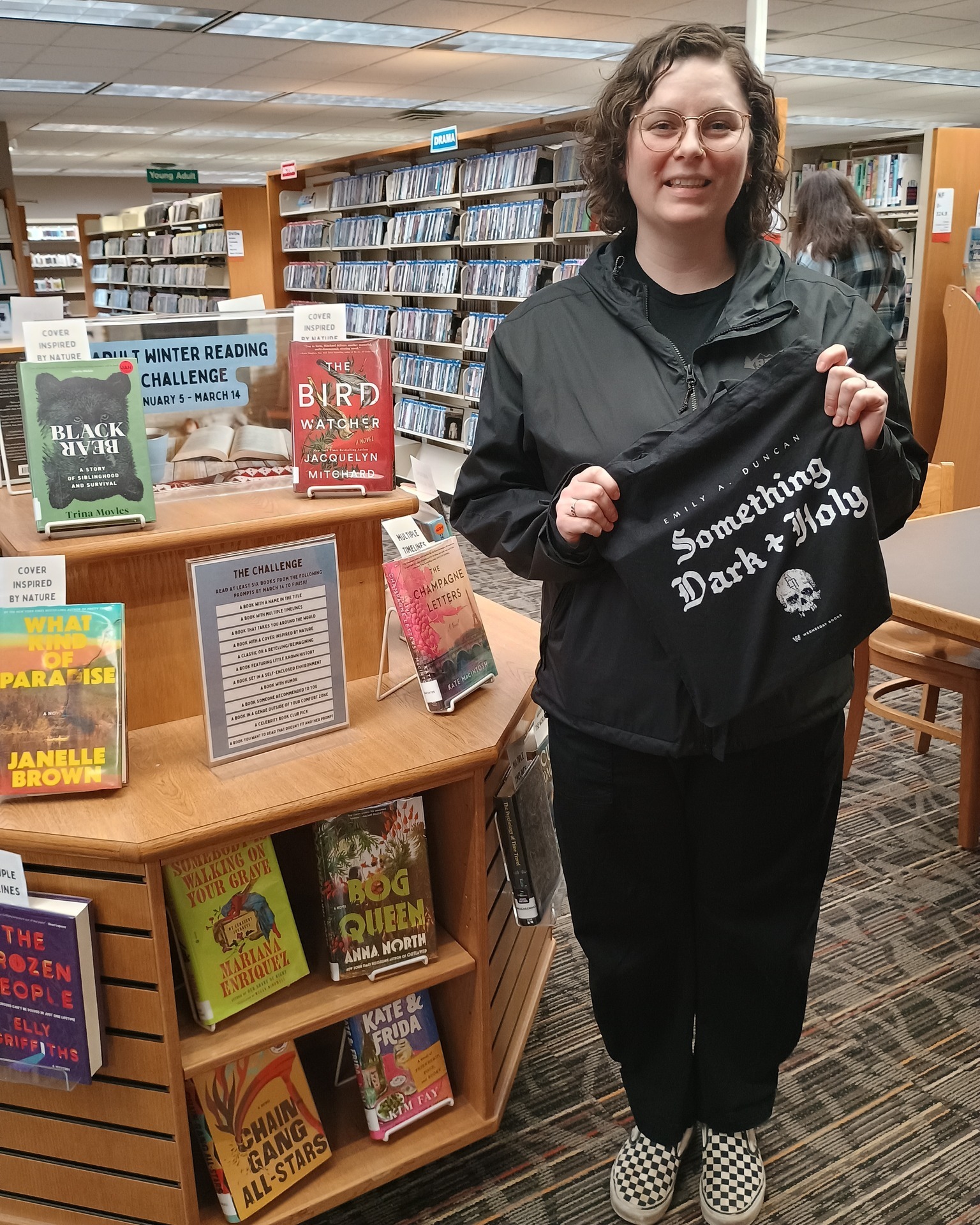 woman stands in library holding a black, nylon bag that reads: Something Dark & Holy