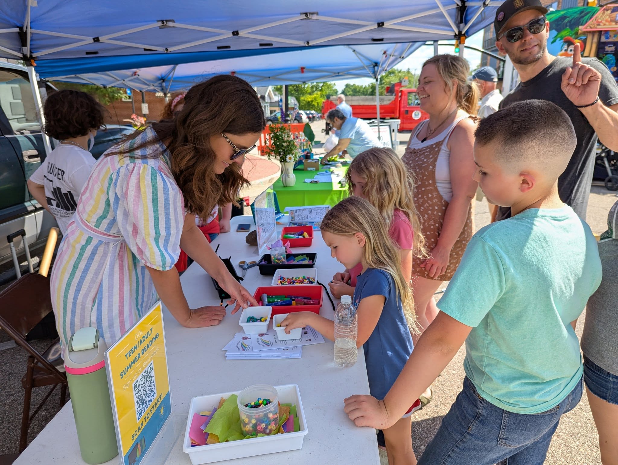 children gather at a table, set up at the DeWitt Farmer's Market, with a library staff who are assisting them in a craft project