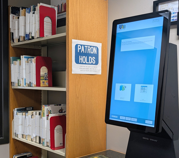 a bookshelf with a sign that reads: Patron Holds standing next to a shelf-checkout station