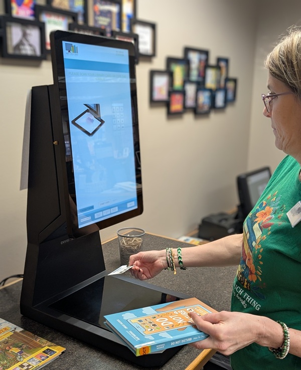 a woman is scanning a library card, while holding a book, at a self-checkout station