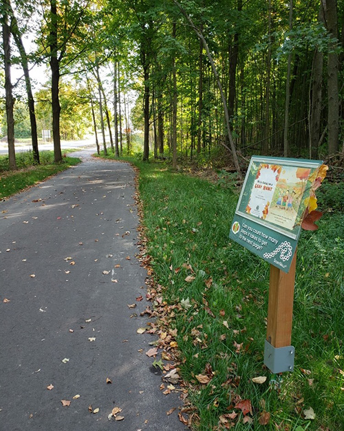 A cement trail going through trees on each side. There is a trail sign on the right hand side.