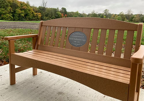 A wooden bench with a green wooded background. The bench has a plaque in memory of Sarah Lago.
