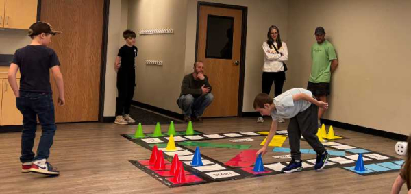 3 adults and 3 kids walk around a Giant Trouble Game on the floor