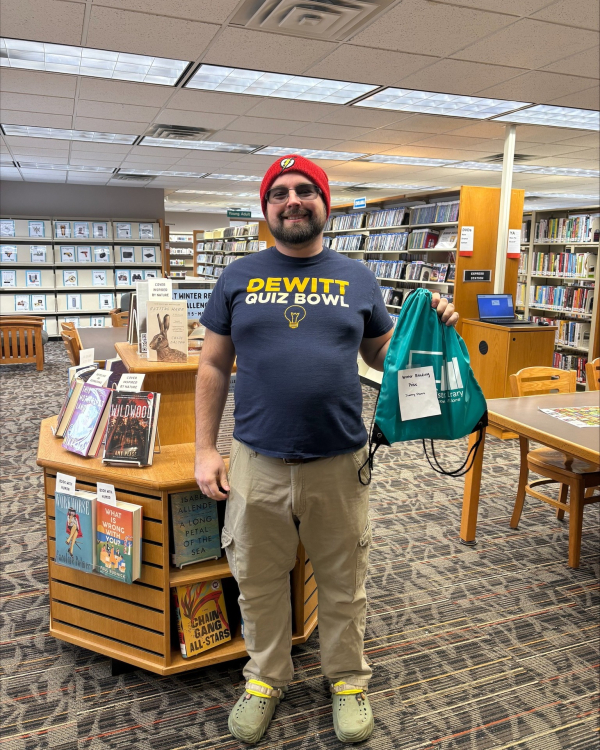 a man wearing a red cap stands in the library wearing a green nylon bag that reads: winter reading prize