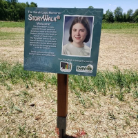 Sign in the middle of a field beside a trail that is a Storywalk trail in memorial of a woman named Sarah Lago.