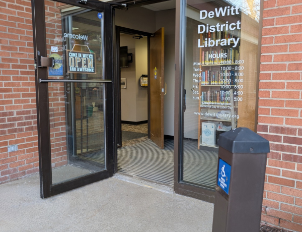 sidewalk that leads to the library entrance doorway, a automatic door post and button in the foreground, an open door, short foyer, and second open door