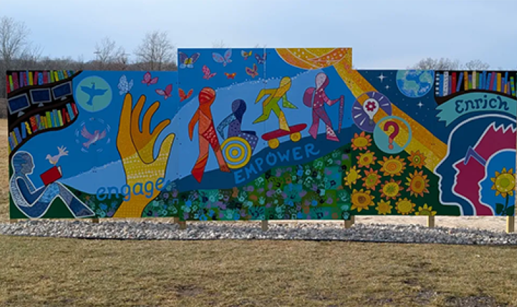 Large six piece canvas mural outside. The mural shows books, a hand reaching out for more, people adventuring outside, and silhouettes of people looking up and enriching their minds.